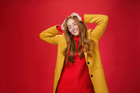 Carefree happy ginger girl carrying away with music and good vibes holding hands on head close eyes and smiling delighted and relaxed having fun, dancing against red background in yellow coatの写真素材