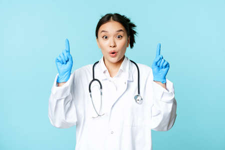 Excited asian medical worker, female doctor pointing fingers up, wearing sterile gloves and uniform, standing over blue backgroundの写真素材