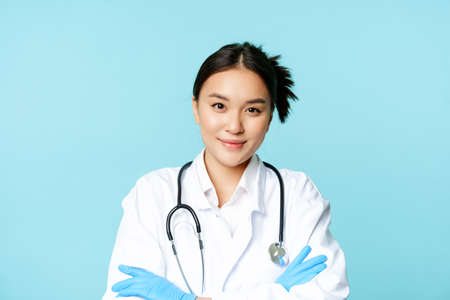 Smiling asian woman doctor, nurse in gloves and clinic uniform, cross arms and looking determined, blue backgroundの写真素材