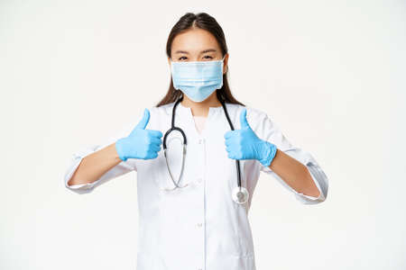 Healthcare and covid-19 preventive measures concept. Asian physician in face mask and rubber gloves, shows thumbs up, stands in uniform over white backgroundの写真素材