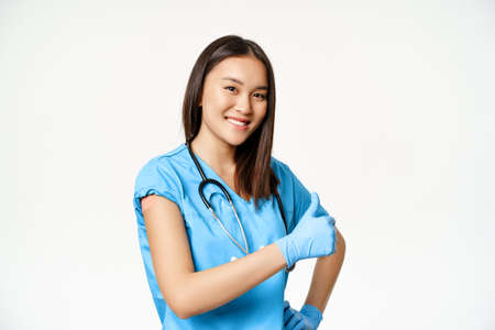 Smiling asian female doctor, nurse in scrubs shows vaccinated hand, flu or covid-19 vaccination campaign, showing thumbs up, white backgroundの写真素材