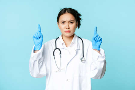 Angry and concerned female doctor, physician pointing fingers up, frowning disappointed, standing in medical uniform over blue backgroundの写真素材