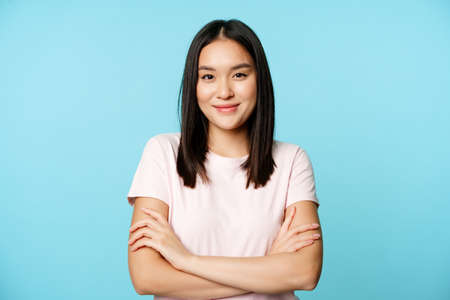 Smiling asian girl in t-shirt, holding arms crossed, ready for action pose, standing over blue backgroundの写真素材