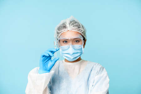 Close up portrait of asian female doctor, nurse in personal protective equipment showing syringe with vaccine, vaccinating from flu or covid, blue backgroundの写真素材