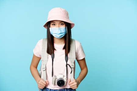 Happy asian girl tourist, standing in medical face mask, holding camera, taking pictures during vacation, sightseeing, explore abroad, standing over blue backgroundの写真素材