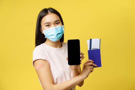 Covid-19 and tourism concept. Smiling asian woman vaccinated, showing smartphone screen, app interface, passport with tickets for vacation, yellow backgroundの写真素材