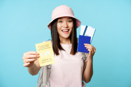 Enthusiastic asian tourist, girl shows covid-19 international vaccination certificate, passport with two tickets, travelling after coronavirus vaccine, blue backgroundの写真素材