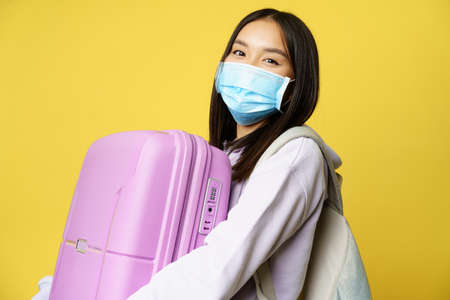 Close up of happy asian girl tourist in medical face mask, holding pink cute suitcase, going on vacation, travelling during pandemic, yellow backgroundの写真素材