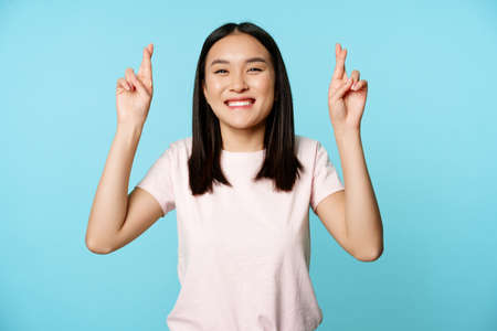 Enthusiastic asian girl wishing, cross fingers for good luck and smiling, peeking at camera hopeful, standing over blue backgroundの写真素材