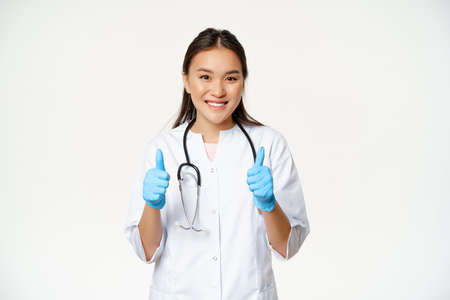 Smiling asian doctor, woman in medical uniform and rubber gloves shows thumbs up, recommending smth to patient, standing over white backgroundの写真素材