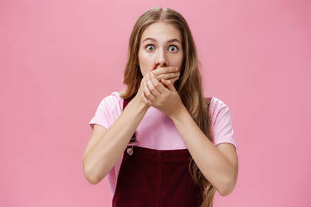 Waist-up shot of shocked speechless pretty young girl in overalls with make-up covering mouth with palms from overreacting on terrible news witnessing accident nervously over pink backgroundの写真素材