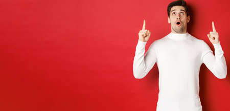 Concept of winter holidays. Portrait of handsome surprised man in white sweater, reacting to new year promo offer, looking and pointing fingers up, standing impressed against red backgroundの写真素材