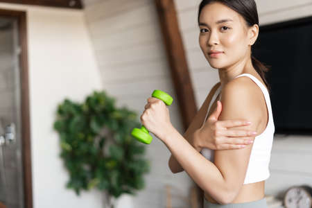 Active and healthy asian girl with fit body doing fitness exercises at home, lifting dumbbells and touching her biceps, workout in living roomの写真素材