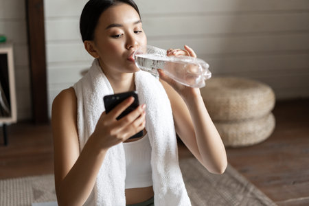 Image of young asian fitness girl with fit body, towel wrapped around neck, looking at mobile phone...