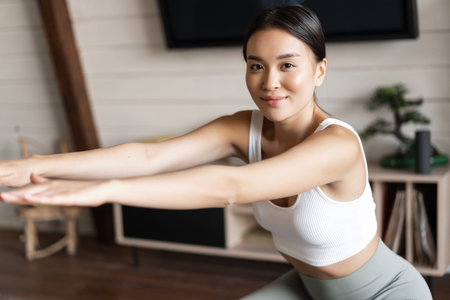 Cute asian fitness girl at home doing workout, squats with stretching elastic rope on her legs, exercising for fit and healthy bodyの写真素材