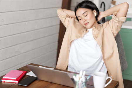 Tired asian girl having break from working, stretching arms after doing homework on laptop, sitting...