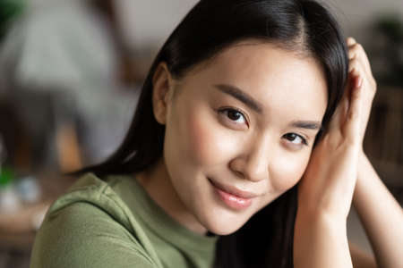 Close up portrait of tender asian girl with glowing natural skin, smiling sensual at camera, sitting at home in cozy clothesの写真素材