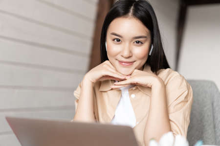 Portrait of asian woman in earphones working on laptop and smiling, looking happy at camera, studying or learning online course on computerの写真素材
