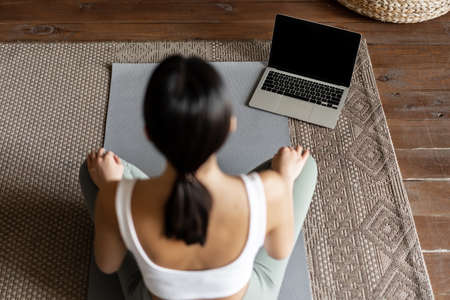 Mindfulness and meditation concept. Rear view of young asian woman meditating at home, following online course on laptop, sitting in living room on floor matの写真素材
