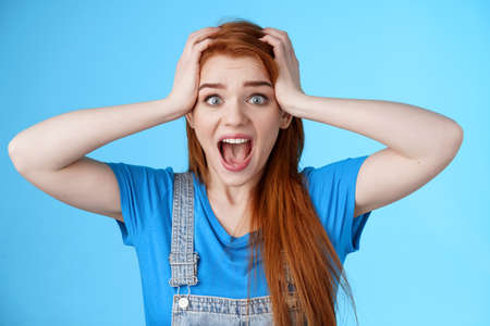 Shocked redhead girl shouting panic, feel pressured deadlines, scared see mess, grab head, scream frightened distressed, stare camera hopeless, anxiously react losing lottery, blue backgroundの写真素材