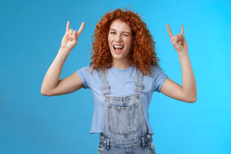 Daring cool sassy redhead ginger girl curly natural hairstyle winking joyfully yelling yeah enjoy awesome party dancing music festival show rock-n-roll heavy metal gesture have fun blue backgroundの写真素材