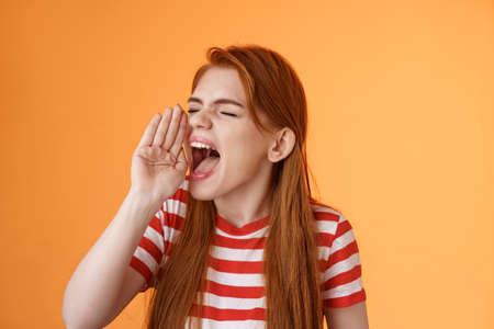 Hey where are you. Cute carefree redhead funny girl hold hand near opened mouth turn left close eyes, yelling out loud, searching someone, calling friend come upstairs, look for person distanceの写真素材