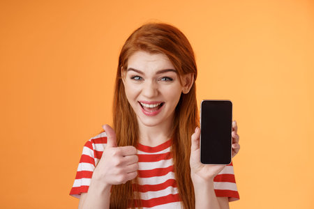 Cheerful assertive redhead woman recommend smartphone app, show phone display, thumb-up like...