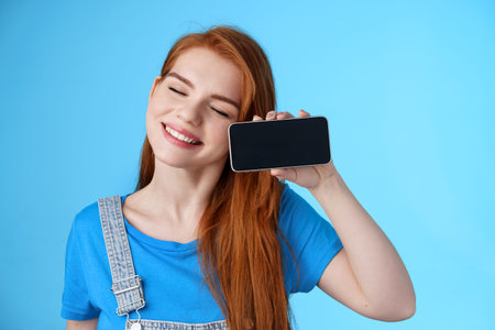 Carefree tender redhead european female tilt head, smiling closed eyes, hold smartphone...