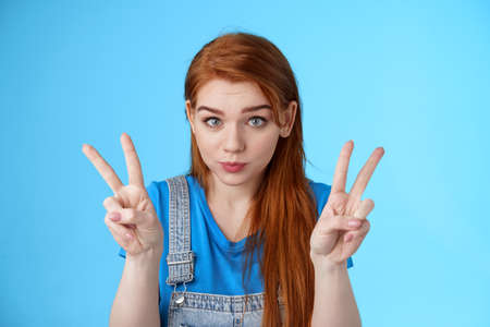 Playful cute silly redhead girlfriend making funny face, show peace victory signs, hold breath, pouting childish, fool around having fun, stand blue background, relaxing entertain siblingsの写真素材