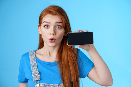 Amazed fascinated excited 20s redhead woman show smartphone screen horizontally, fold lips whistling amused wondered, check out cool phone app, stand blue background curiousの写真素材