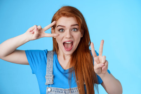 Cute excited redhead enthusiastic girl smiling, look joyful show peace, victory signs near face, optimistic lucky attitude, believe win, stand blue background positive, having fun enjoy vacationの写真素材