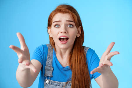 Close-up uneasy disappointed cute gloomy redhead girl, raise hands dismay full dibelief, frowning gasping upset, losing competition, receive bad results, stand blue background unhappyの写真素材