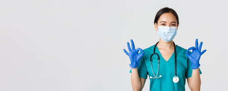 Covid-19, coronavirus disease, healthcare workers concept. Confident smiling asian female doctor, nurse in medical mask and gloves, show okay gesture in approval, white backgroundの写真素材