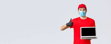 Customer support, covid-19 delivery packages, online orders processing concept. Cheerful courier in red uniform, medical face mask and gloves recommend webpage, show laptop screen and thumb-upの写真素材