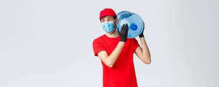 Takeaway, food and groceries delivery, covid-19 contactless orders concept. Carefree courier in red uniform cap, face mask and gloves, hold bottled water on shoulder, look away with smileの写真素材