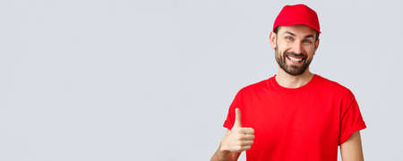 Online shopping, delivery during quarantine and takeaway concept. Friendly, cheerful courier in red cap and t-shirt uniform, encourage make internet orders, thumb-up in approval or recommendationの写真素材