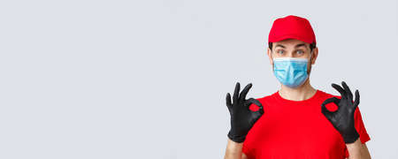 Contactless delivering, covid-19 and shopping concept. Cheerful, pleased courier in red uniform, cap and medical mask with gloves show okay, approval or guarantee gesture, recommend serviceの写真素材