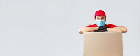 Packages and parcels delivery, covid-19 quarantine and transfer orders. Young courier in red uniform cap, face mask and gloves, leaning on box to deliver, shipping your orders, grey backgroundの写真素材