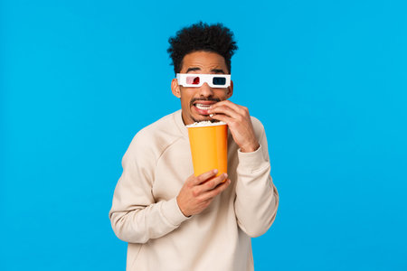 So scarry but interesting. Enertained handsome funny african american guy watching horror movie in cinema, eating popcorn and cringe looking screen, standing blue background tensedの写真素材