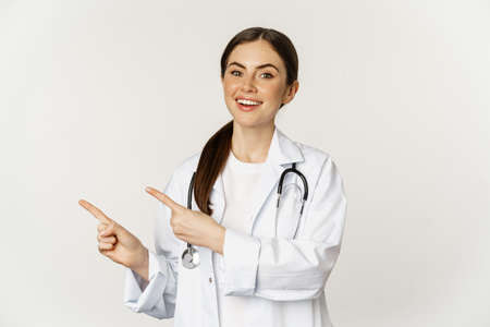 Portrait of smiling young woman doctor, healthcare medical worker, pointing fingers left, showing clinic promo, banner, standing over white backgroundの写真素材