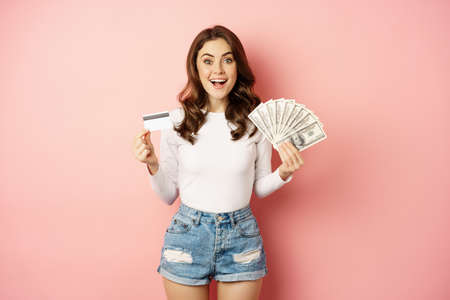 Credit, money and loans concept. Happy beautiful girl holding credit card and cash, looking satisfied, thinking of shopping, standing over pink backgroundの写真素材