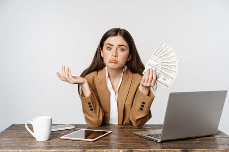 Sad businesswoman sitting in office with laptop, holding money and looking disappointed in income,...