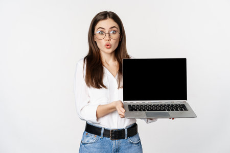 Portrait of cute girl in glasses, student showing laptop screen with amazed face, standing over white backgroundの写真素材