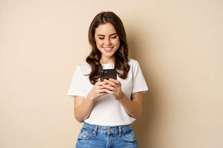 Young modern girl chatting on app, using smartphone app and smiling, standing over beige backgroundの写真素材