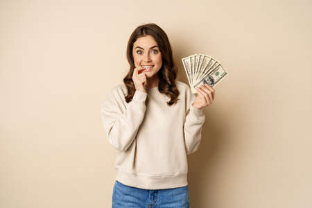 Excited brunette woman holding money and biting finger with yearning face, shopaholic with cash, standing over beige backgroundの写真素材