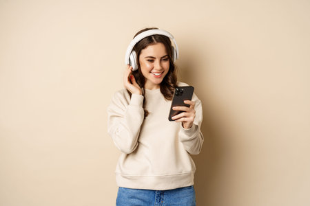 Beautiful young woman looking at video on smartphone, listening music in headphones, standing in...