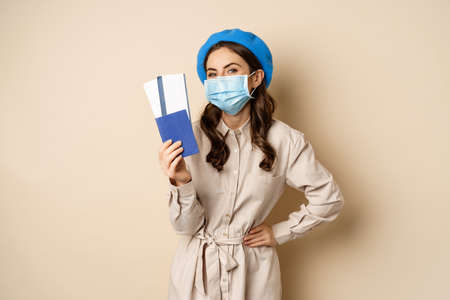pandemic and traveling concept. Young woman tourist showing her passport and tickets abroad, wearing face mask from coronavirus, posing over beige backgroundの写真素材