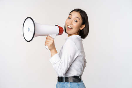 Beautiful young asian woman talking in megaphone, screams in speakerphone and smiling, making announcement, shout out information, standing over white backgroundの写真素材