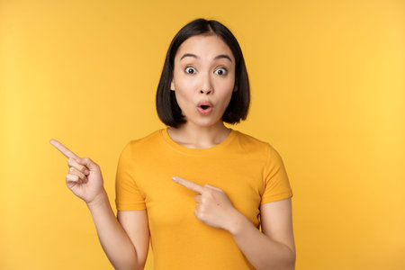 Smiling asian woman pointing fingers left, showing advertisement on empty copy space, standing over...