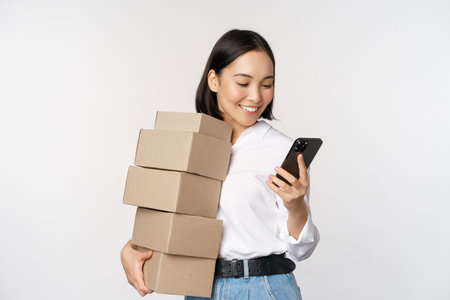 Image of young asian woman holding boxes, customer orders and looking at mobile phone, standing over...